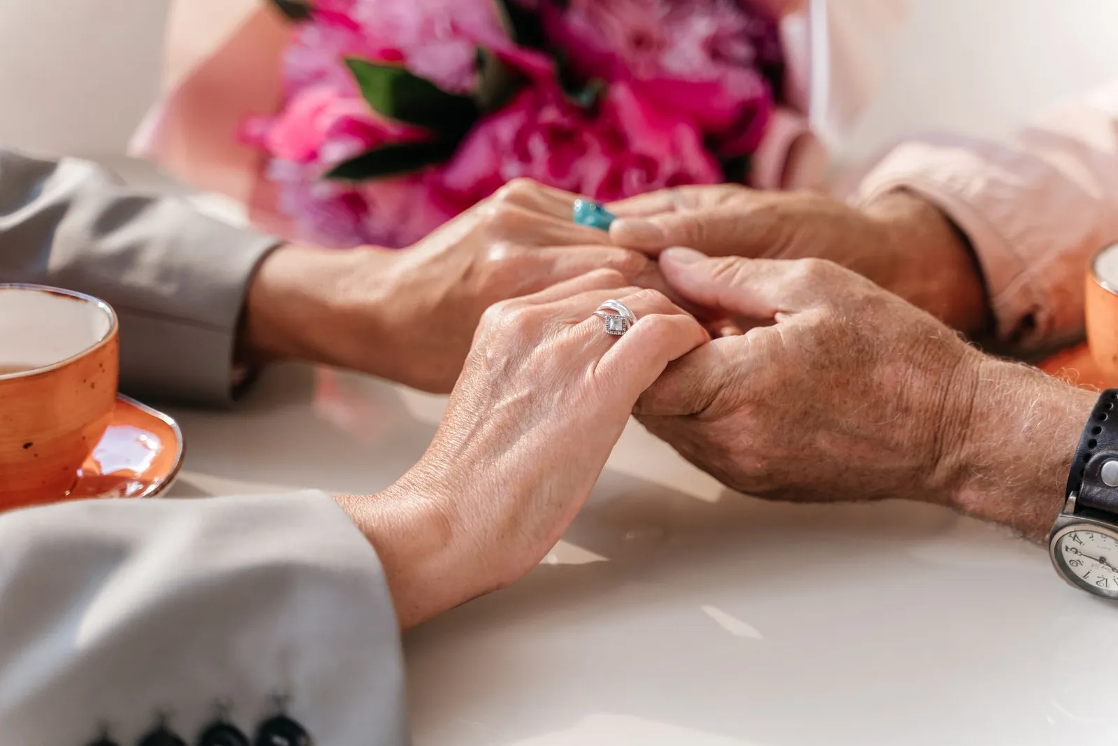 An older couple with hands clasped, wedding rings and flowers
